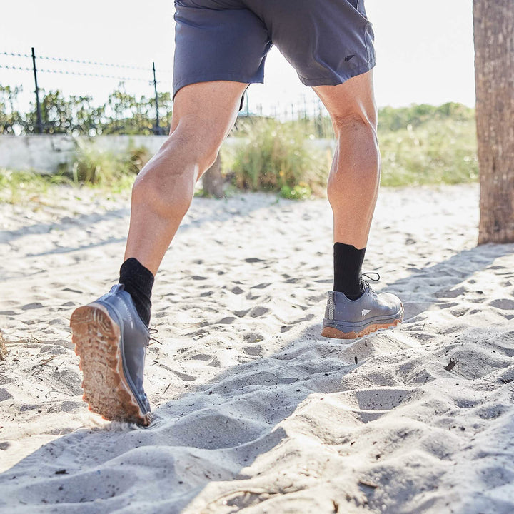 A person wearing GORUCK Mackall shoes in Forged Iron, Chiseled Stone, and Gum colors, along with black socks, is jogging on a sandy beach. They are dressed in gray shorts with a tree trunk and grassy area in the background under sunny skies.