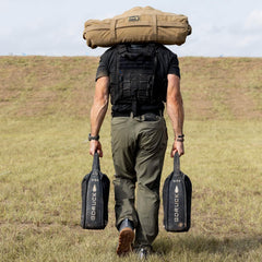 Man wearing tactical vest carrying GORUCK sandbags and forged iron bag over shoulders on grassy field