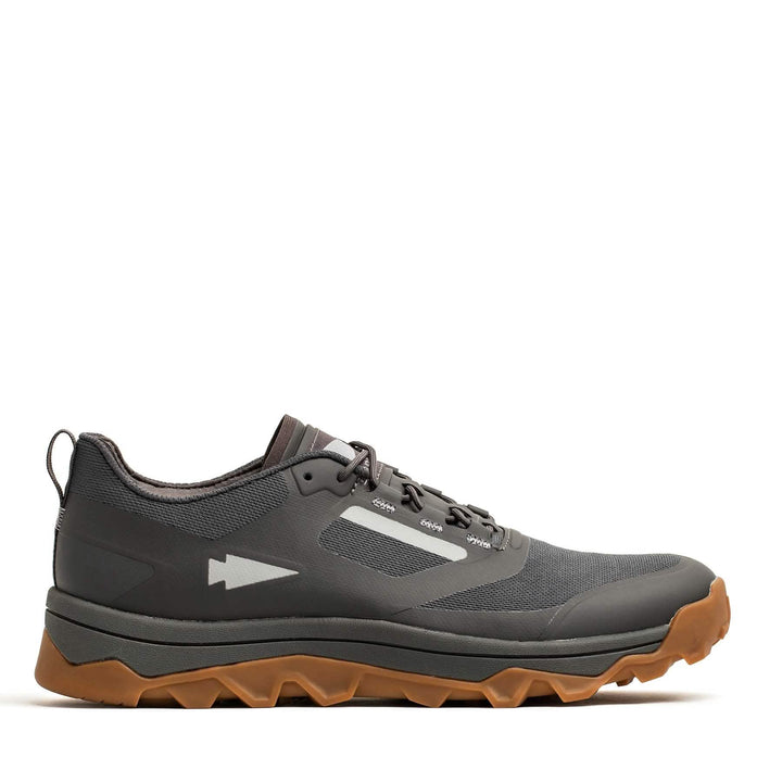 The GORUCK Mackall in Forged Iron, Chiseled Stone, and Gum is a trail shoe crafted for outdoor activities. It boasts a minimalist design with a gray and black color scheme, complemented by a brown sole. The all-terrain textured outsole ensures excellent grip, and a small arrow symbol adorns the side. The background is plain white.