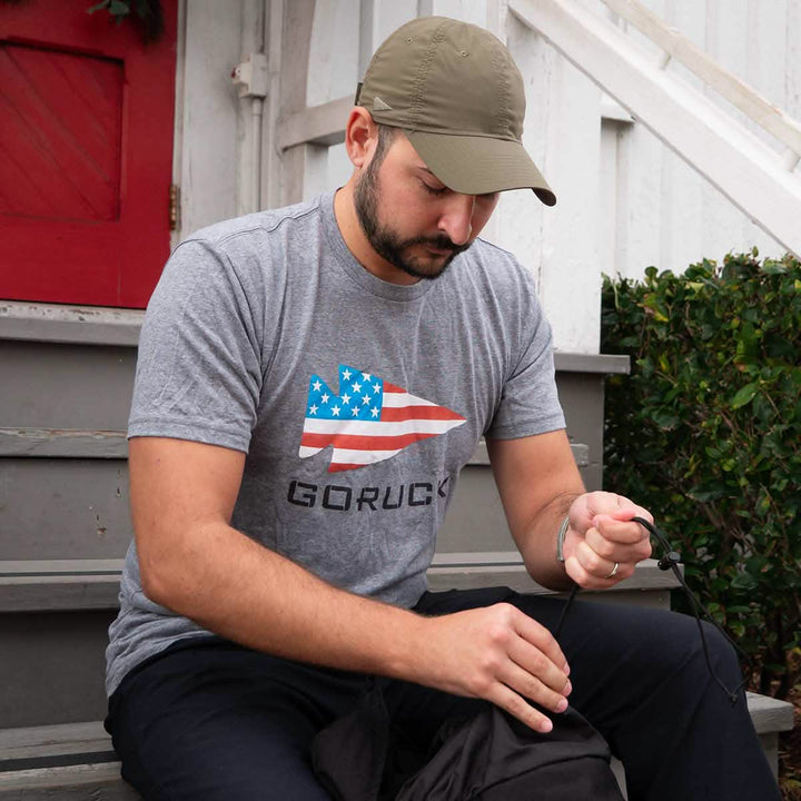 Man wearing a gray GORUCK USA Tee - Tri-Blend and olive cap sits on steps, threading cord through a black bag—a nod to Special Forces precision.
