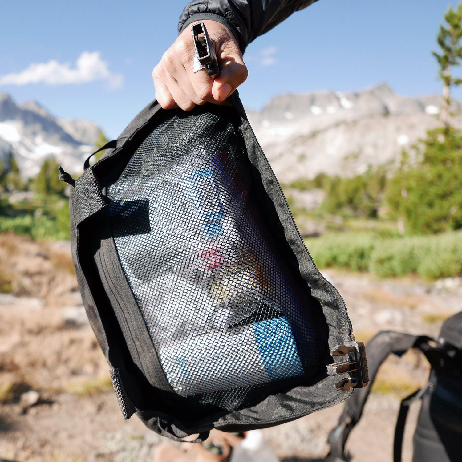 A hand holds the GR MONSTER Bundle - PREORDER in a mesh bag with gear inside, outdoors in a mountainous landscape with trees and rocks—perfect for stowing alongside your trusted GORUCK rucksack.
