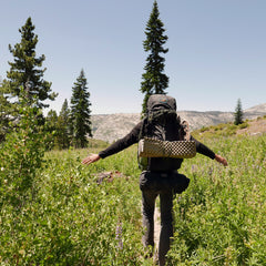A hiker carrying the GR MONSTER Bundle - PREORDER rucksack walks through green bushes on a mountain trail with arms outstretched, trees and hills in the background.