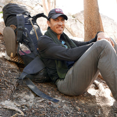 A man in outdoor gear sits on a slope with the GR MONSTER Bundle - PREORDER rucksack from GORUCK, smiling at the camera in a forested area.