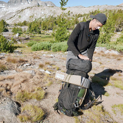 A person adjusts the GR MONSTER Bundle - PREORDER rucksack outdoors, surrounded by mountains, trees, and rocky terrain in sunlight.