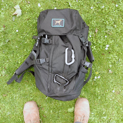 The GR MONSTER Rucksack - PREORDER in black, featuring a dog patch and carabiner, is shown lying on the grass near tan boots.