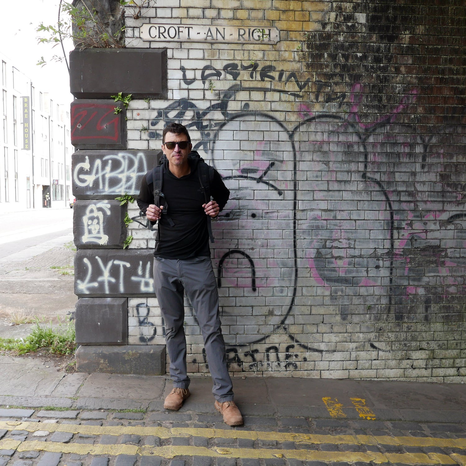 A man in sunglasses stands by a graffiti-covered brick wall under a bridge, dressed in casual clothes and carrying the GR MONSTER Rucksack - PREORDER on his back.