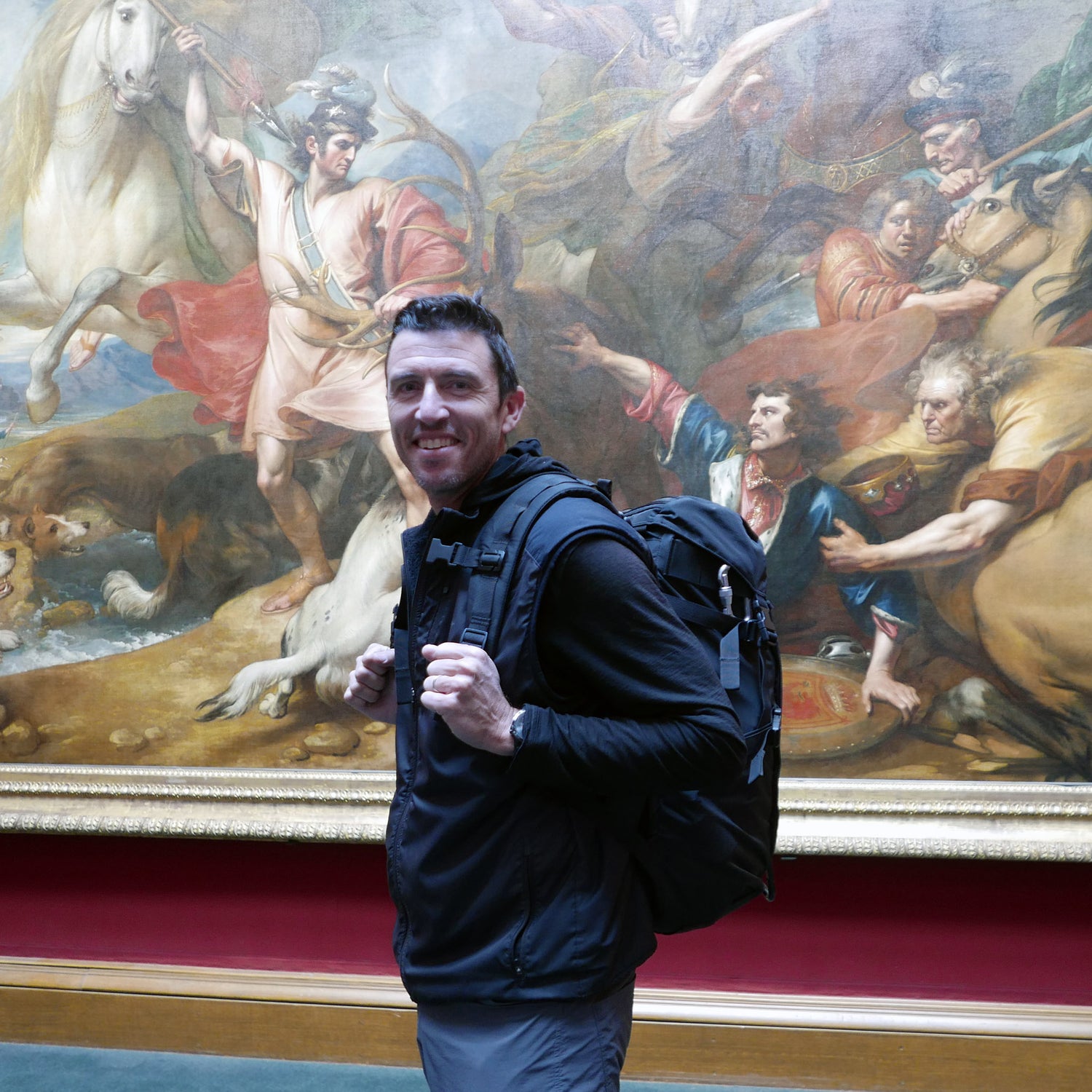 A man with a GR MONSTER Rucksack - PREORDER smiles in front of a large classical painting in a museum.