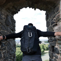 A person with a large black GR MONSTER Rucksack - PREORDER stands in a stone arch, overlooking trees and a distant city.