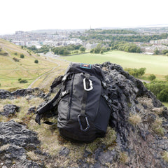 A black GR MONSTER Rucksack - PREORDER with carabiners sits on rocky ground, overlooking a sprawling city and green fields below.