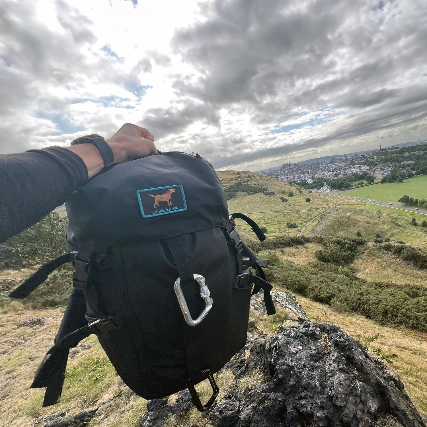 A hand holds the GR MONSTER Rucksack - PREORDER in black, featuring a dog patch, with a city and green hills visible under cloudy skies.