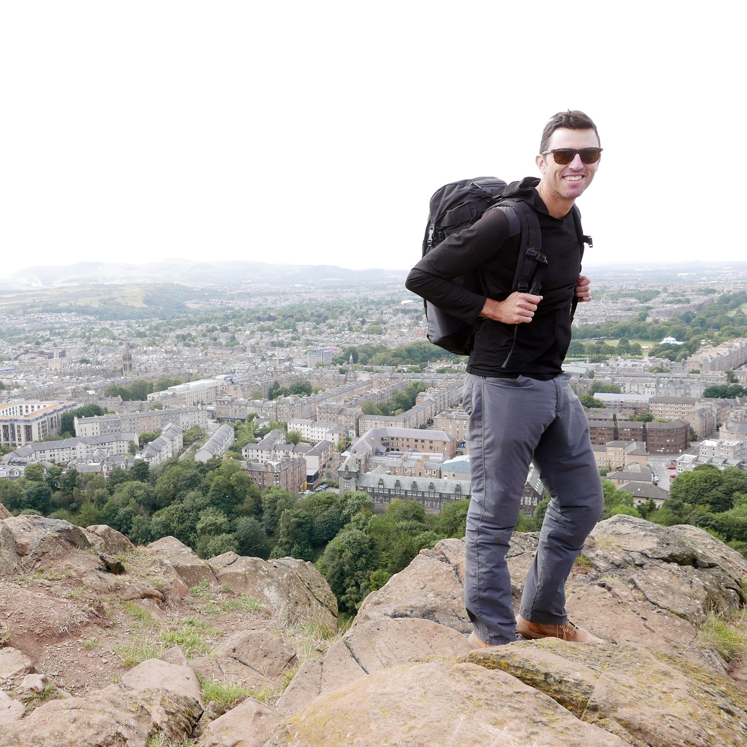 A man wearing sunglasses stands on a rocky hill overlooking a cityscape and greenery, carrying the GR MONSTER Rucksack - PREORDER.