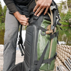 A person securing a GR MONSTER Rucksack - PREORDER in green and black near a body of water in an outdoor setting.