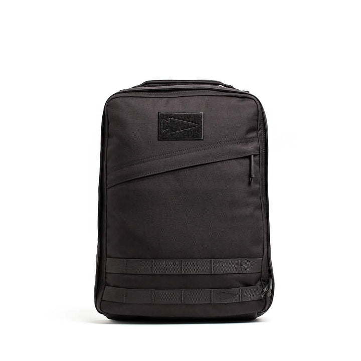 The GR0 - Cordura by GORUCK is a black rectangular compact backpack with a sleek, minimalist everyday carry design. It includes a front zippered pocket, a stitched arrow logo patch, and horizontal strap detailing at the bottom. The bag stands upright against a white background.