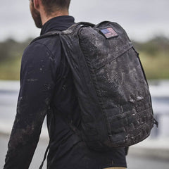 A person carrying a GR1 USA - Cordura backpack by GORUCK, complete with a US flag patch, strides through the outdoors, looking ready for a GORUCK Challenge.