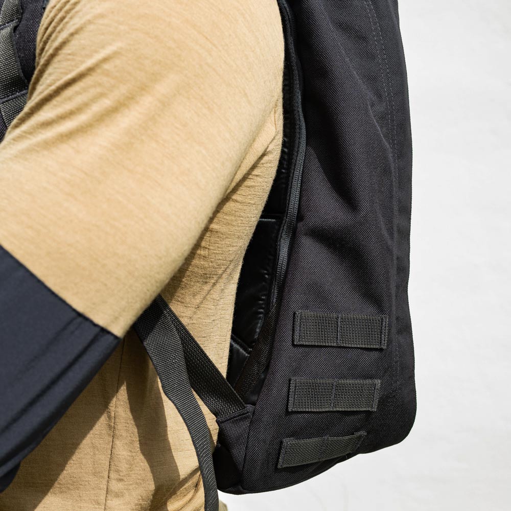 Person wearing a tan shirt and a black backpack with webbing straps on the side, against a light background.