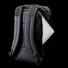 A black GORUCK GR1 USA - Heritage rucksack with padded straps is displayed against a black background. It has a side zipper pocket partially open, revealing the edge of a laptop.