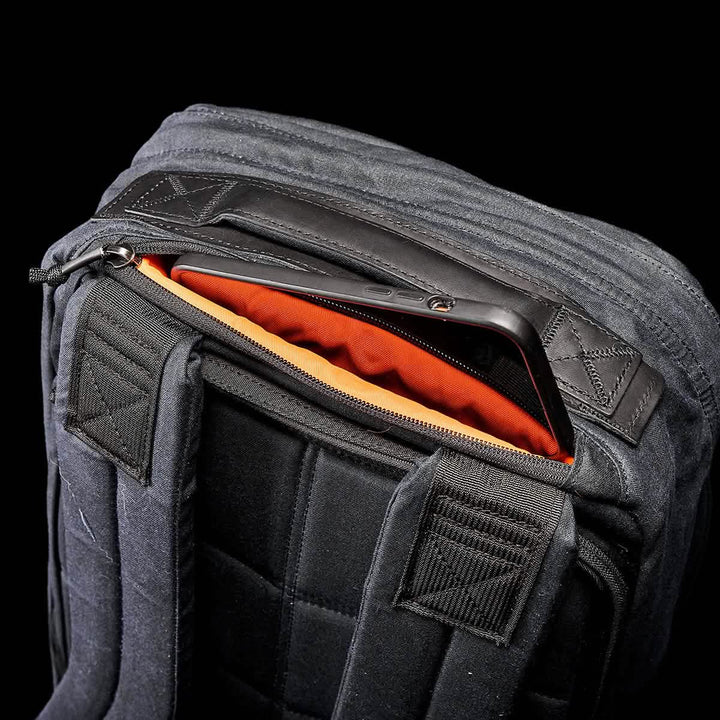 Close-up of the GR1 USA - Heritage black backpack by GORUCK, showcasing a partially open top zipper that reveals an orange interior with a tablet or laptop inside. The retro canvas rucksack features padded straps and a sleek, quilted design, set against a black background.