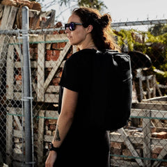 A person with sunglasses and a tattoo on their arm stands outdoors, wearing a black shirt and a GORUCK GR1 USA - Heritage backpack. Near a chain-link fence and woodpile, sunlight casts a natural, rugged atmosphere, emphasizing the durable style of the Waxed Duck Canvas material.