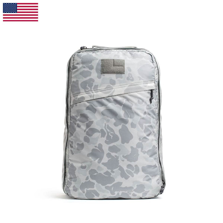 The GR1 USA - Ripstop ROBIC® by GORUCK is featured on a white background with an American flag in the top left corner. This ultra-lightweight, gray camouflage backpack includes a zippered front pocket, a carrying handle at the top, and showcases its water-resistant design.