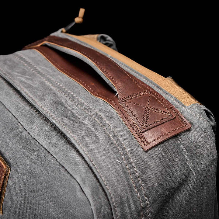 Close-up of the GORUCK GR1 USA - Heritage, a gray rucksack showcasing a brown leather handle with intricate stitching. The texture of its waxed duck canvas and leather contrasts beautifully against the black background, highlighting the materials.
