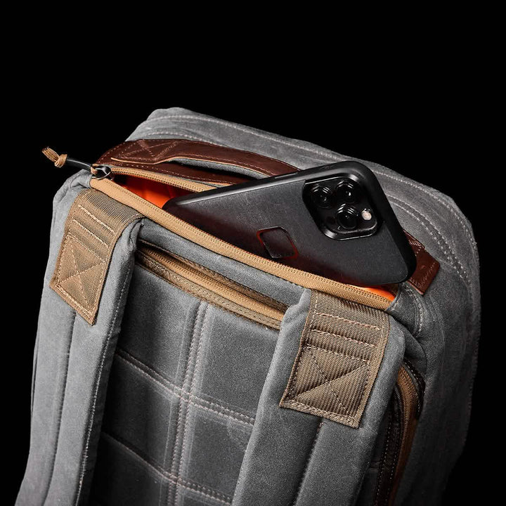 The GR1 USA - Heritage by GORUCK in gray features tan straps and is partially open, revealing a black smartphone inside the front compartment. The rucksack showcases quilted stitching with a brown leather accent at the top, all set against a black background.