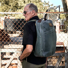 A man wearing sunglasses and a black shirt carries a large green GR1 USA - Heritage rucksack by GORUCK. He is standing in front of a chain-link fence, with industrial equipment and wooden pallets in the background.