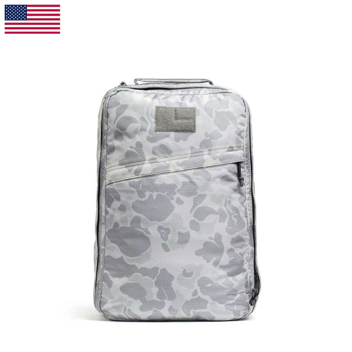 The GR1 USA - Ripstop ROBIC® by GORUCK is a gray camouflage-patterned rucksack featuring a rectangular patch on the front and a diagonal zipper pocket. Its ultra-lightweight design and the U.S. flag displayed in the top left corner make it both stylish and water-resistant for all your adventures.
