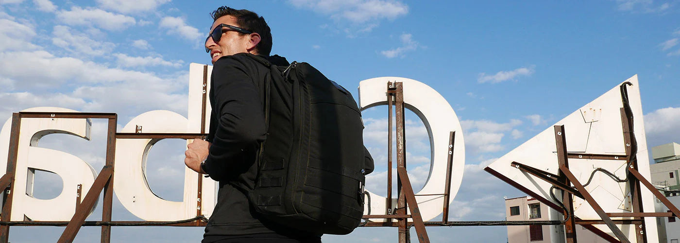 Man wearing sunglasses and a black backpack stands in front of a large rooftop sign against a blue sky.