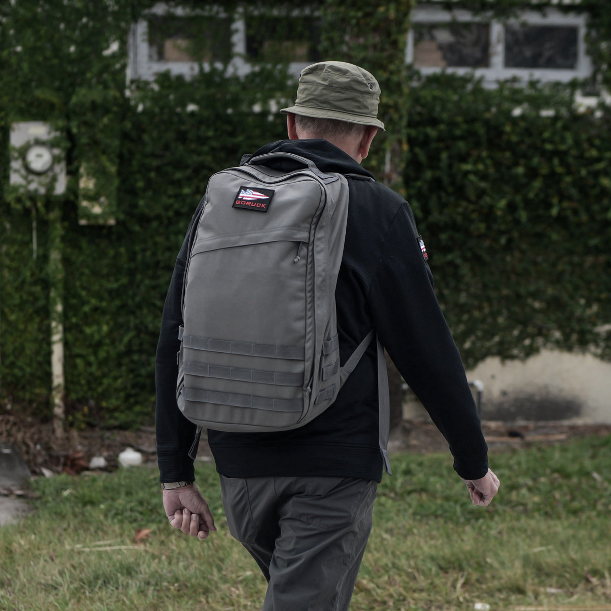 A man in a green hat and gray GR1 USA - Cordura rucksack, an everyday carry backpack, walks outside near a vine-covered building.