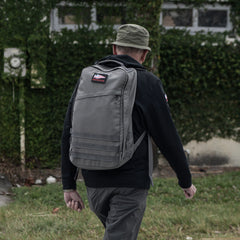 A man in a green hat and gray GR1 USA - Cordura rucksack, an everyday carry backpack, walks outside near a vine-covered building.