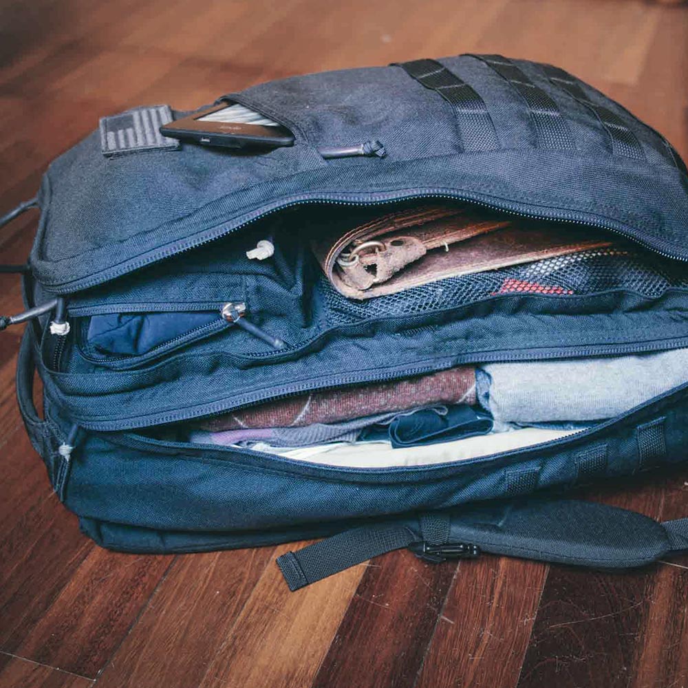 A black backpack unzipped, showing clothes, a leather item, and a phone on top, on a wooden floor.