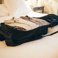 A GR2 travel bag from GORUCK, in sleek black, sits open on a bed, expertly packed with folded clothes. The room is softly lit, and the bed is adorned with white linens. In the background, there is a side table featuring a small lamp.