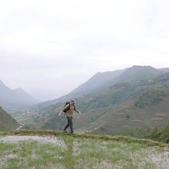 A person wearing a hat and the GORUCK GR2 backpack, celebrated for its Special Forces quality, walks across a grassy hill. Terraced fields and mountains stretch beneath a cloudy sky. With its perfect carry-on compliant size, the backpack is ready for any adventure.