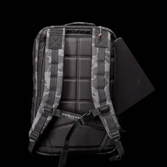 A GR2 backpack by GORUCK, reminiscent of Special Forces gear, features padded shoulder straps and grid-like back padding. A laptop peeks from its compartment, while a small GORUCK label subtly brands the ruck against the solid black background.