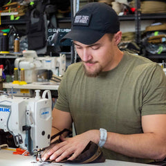 A man, dressed in a cap and green shirt, sews fabric on an industrial sewing machine in a workshop as he carefully assembles the Slick Short Sleeve Tee - Tri-BlendX, ensuring every stitch meets the highest standards of quality.