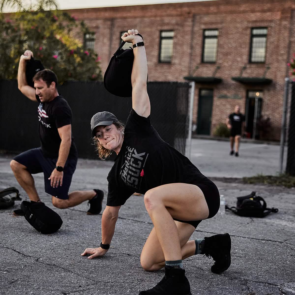 Two individuals are working out outside on a paved surface. One is kneeling and lifting a sandbag overhead, while the other mimics the movement. They are both wearing shorts and sweat-wicking T-shirts, and one of them sports a GORUCK Performance TAC Hat made with TOUGHDRY fabric. In the background, there's a brick building and fence, with another person seen in the distance.