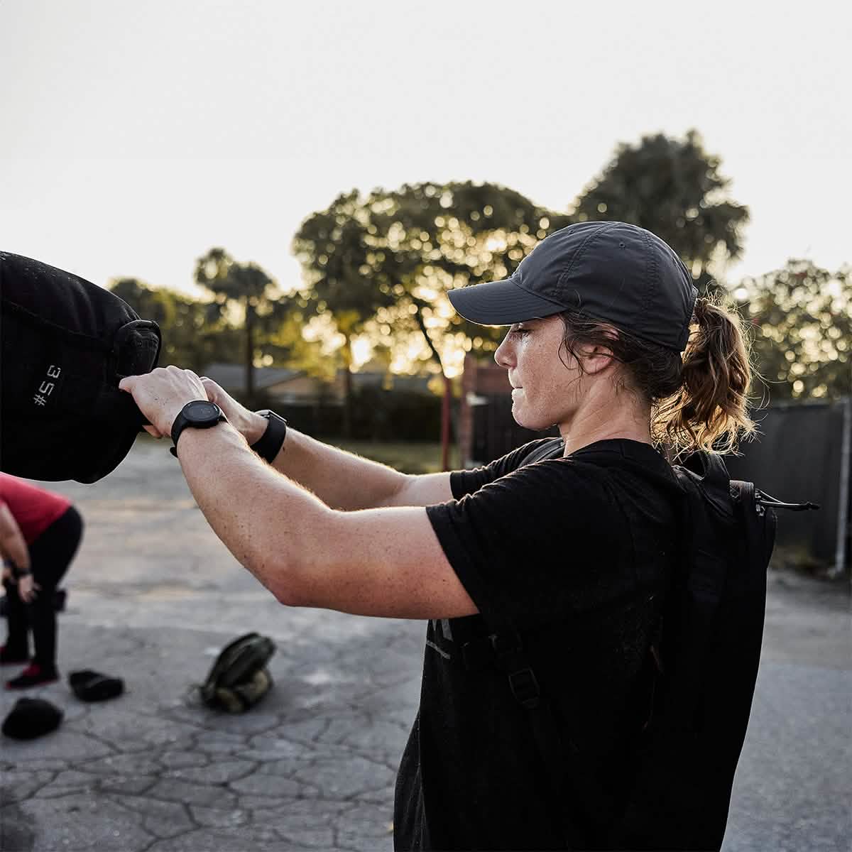 A person wearing the GORUCK Performance TAC Hat - Slick made with TOUGHDRY material and a black shirt is exercising outdoors, lifting a weighted sandbag. In the background, another individual bends over. The scene unfolds on a paved area dotted with trees under a clear sky, perfect for highlighting the effectiveness of sweat-wicking gear in action.