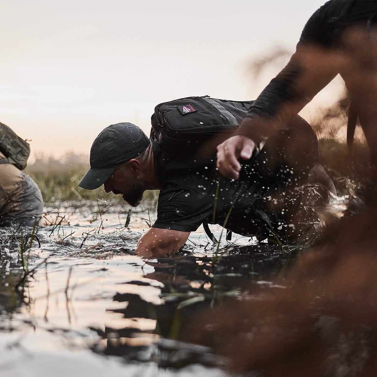 A person outfitted in outdoor gear, including a GORUCK Performance TAC Hat - Slick made with TOUGHDRY fabric and a ToughDry® backpack, navigates through water and grass. This unfolds in a natural setting during dusk or dawn, with another individual partially visible in the background. Water splashes around them glisten under the soft light.