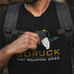 A person wearing a GORUCK t-shirt from the GRT Reunion 2020, crafted from durable 1000D CORDURA and featuring gold and blue lettering with a graphic, adjusts the chest strap of their black Ruck Plate Carrier 3.0. Outdoors, they rely on the backpack's ergonomic lumbar support for comfort.
