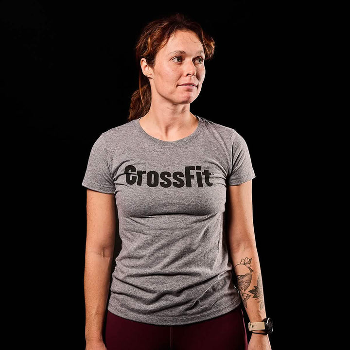 A person wearing a Women's CrossFit Tee - Tri-Blend by GORUCK in gray, featuring a USA print, stands against a black background.