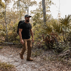 A man wearing a black ToughDry® t-shirt, brown pants, and a GORUCK Performance TAC Hat - Slick is hiking on a dirt trail in the forest. With his black backpack in tow, he's encircled by dense green foliage and trees. The sun filters softly through the branches as he enjoys the sweat-wicking comfort of his attire on his journey.