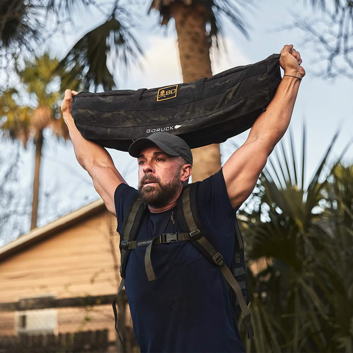 A person wearing a GORUCK Performance TAC Hat - Slick, crafted with TOUGHDRY fabric, and a black T-shirt lifts a heavy sandbag over their shoulders while working out outdoors. The sweat-wicking properties keep them cool as palm trees and a house frame the background.