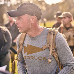 A man wears the Men's GORUCK Spearhead Long Sleeve - Tri-BlendX shirt in gray and a cap, carrying a backpack outdoors in sunlight, with people visible in the background.