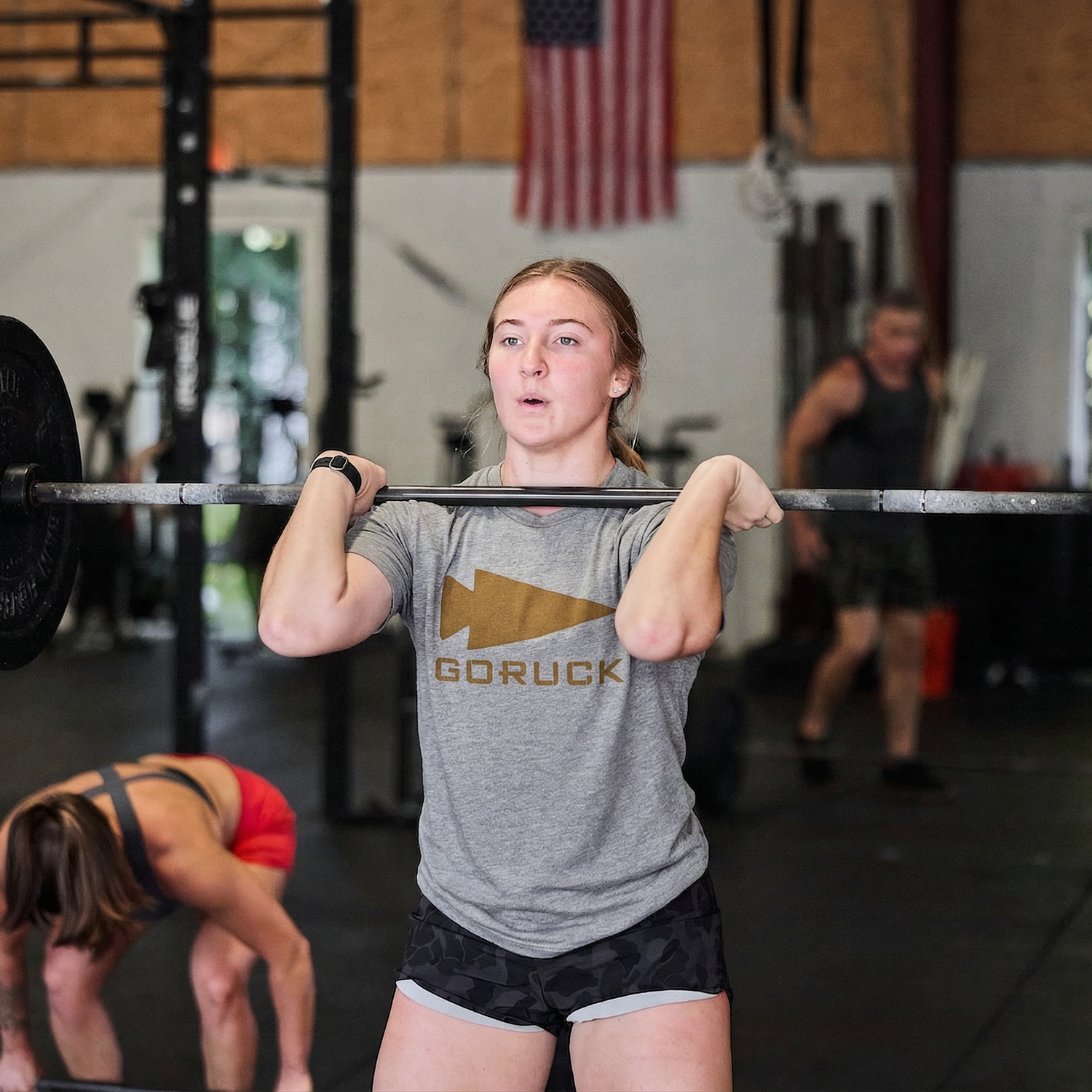A young woman lifts a barbell in the gym wearing the GORUCK Spearhead Tee - Tri-Blend, with others training nearby and an American flag plus a GORUCK spearhead banner in the background.
