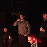 Group of men in GORUCK gear enjoying drinks around a campfire at night outdoors.