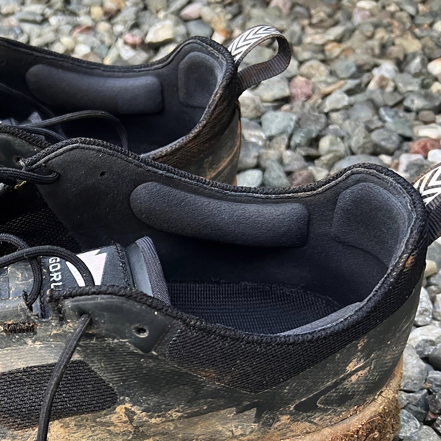 Close-up of two black muddy trail shoes with thick padding, resting on gravel.