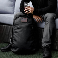 A person sitting on a couch outdoors, holding a glass of beer in one hand and resting the other on a GR1 USA - Heritage rucksack by GORUCK placed on the ground. The person is wearing dark clothing, creating a relaxed atmosphere.