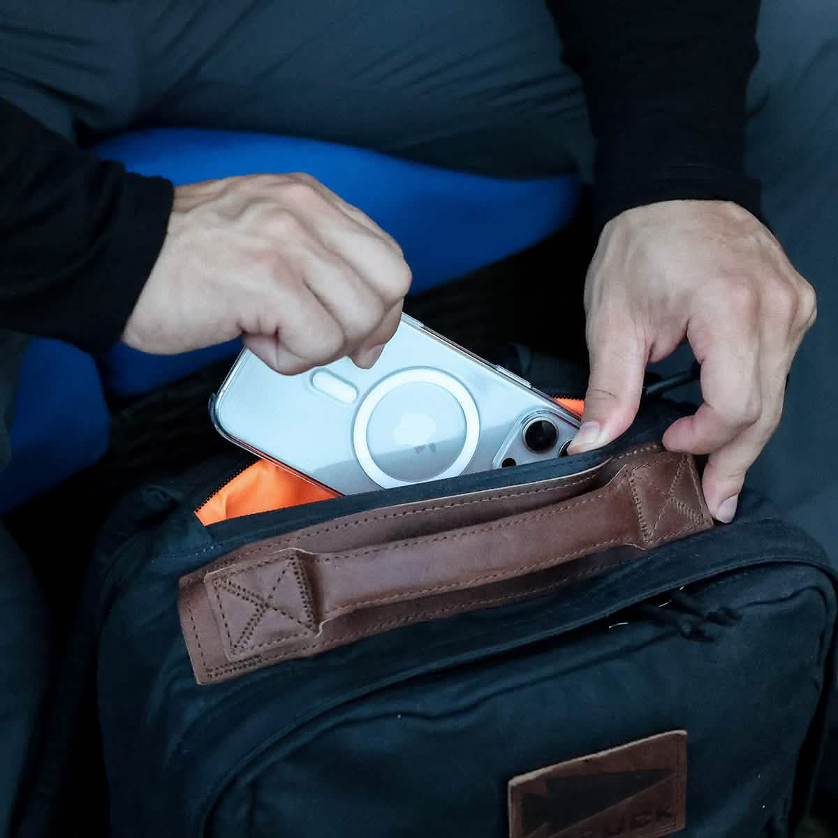 A person places a smartphone with a clear case into the GORUCK GR1 USA - Heritage, a black backpack featuring brown leather accents. The interior of this chic waxed duck canvas rucksack is bright orange. They're wearing a long-sleeve black shirt that effortlessly blends style and functionality.