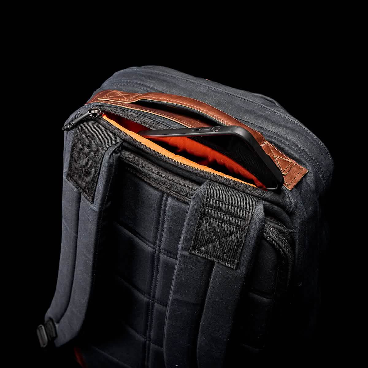 A GR1 USA - Heritage by GORUCK, featuring brown leather accents and partially unzipped to reveal a smartphone inside, highlights its vibrant orange lining. Crafted from waxed duck canvas, this black backpack rests elegantly against a black background, blending classic style with modern utility.