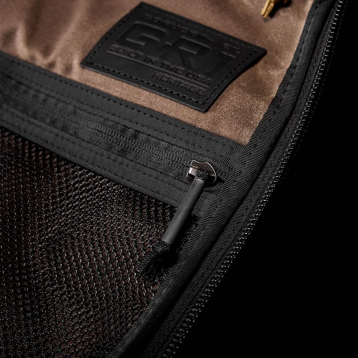Close-up of a GR1 USA - Heritage rucksack by GORUCK, featuring brown fabric with black mesh pockets, a zipper, and a metal pull tab. A tag states "GR1" and "Built in the USA." The texture and stitching details resemble classic retro canvas designs.